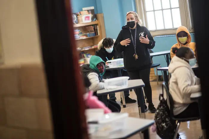 Masked students sit at desks with a teacher, wearing a mask, stands in the classroom; the photographer is looking in at the classroom from the hallway
